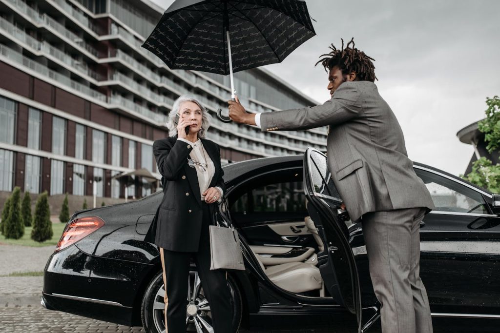 Man holds umbrella for businesswoman on phone beside luxury car and building.